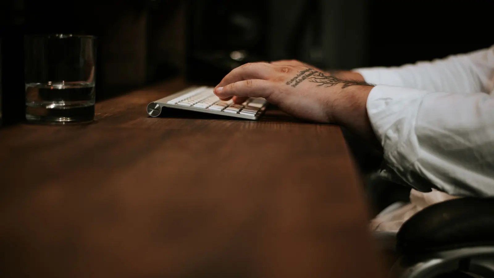 Mão apoiada em um teclado branco sobre uma mesa de madeira escura, com um copo de vidro ao lado e luz baixa de ambiente